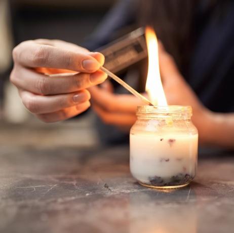 реколта анонимна жена с горящ кибрит осветяване ароматна свещ в стъклен буркан поставена на мраморна маса у дома