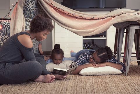 майка седи до крепост от одеяла с двете си дъщери и чете история от книга