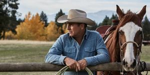 Кевин Костнър в Йелоустоун до кон, облегнат на ограда с въже в ръце, облечен в избеляла синя дънкова риза и бежова каубойска шапка