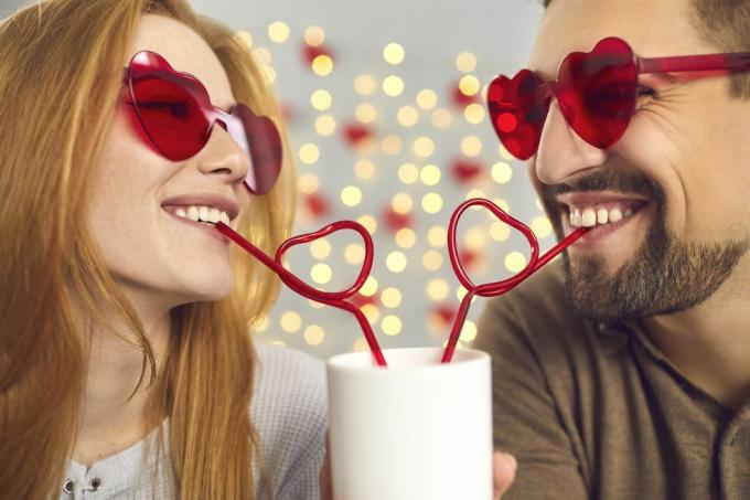 портрет в близък план щастливи млад мъж и влюбена жена в розови слънчеви очила, отпиващи от една чаша през сламки във формата на сърце, наслаждавайки се на сладък забавен момент на двойка на забавна среща в деня на Свети Валентин