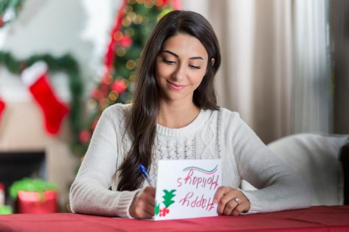 усмихната млада жена седи на маса в декорираната си всекидневна и гледа съсредоточено надолу, докато пише писмо до приятел за Коледа