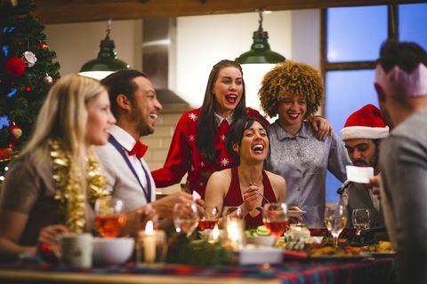 група приятели се смеят, докато седят около маса, ядат и пият по Коледа, един мъж чете виц от коледен крекер, носени са шапки и коледни джъмпери